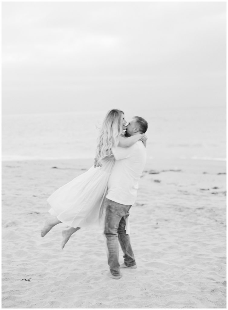black and white portrait of husband twirling his wife in the air at the beach