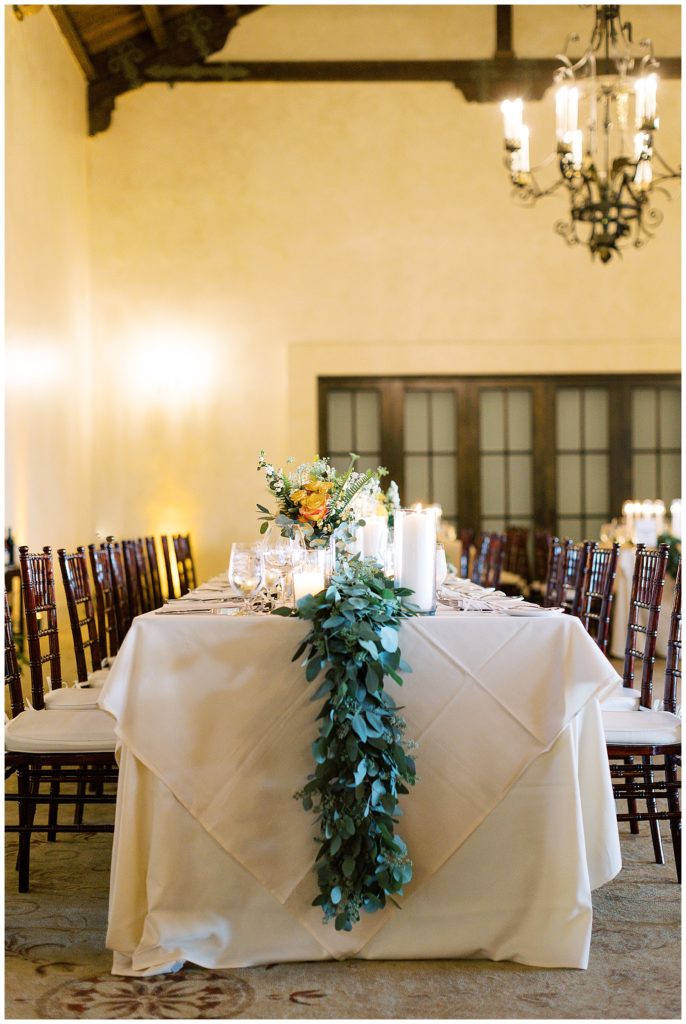 MPCC luxury wedding dining table set up with lush green florals, tall candles, wooden chairs, and an antique chandelier