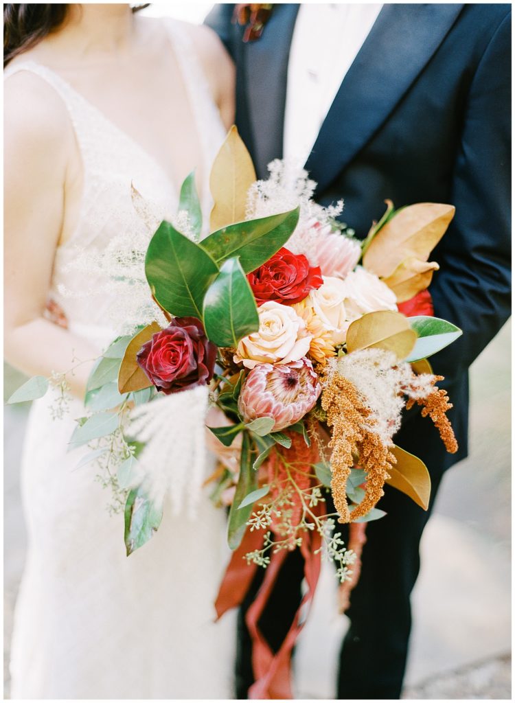 photo of the married couple from the shoulders down with a focus on the bride's red, pink, and orange bouquet by Big Sur Flowers by film photographer AGS Photo Art