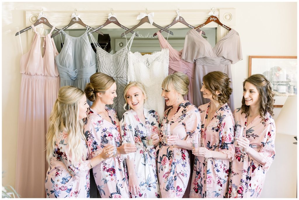 Six women in blush pink floral robes hold champagne glasses in front of different shades of BHLDN wedding dresses. Anthropologie inspired picture as they all toast and smile. 