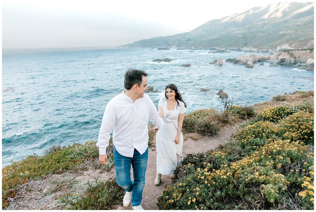 couple walking along the cliffside lined with yellow flowers on one side and rolling blue ocean waves on the other side