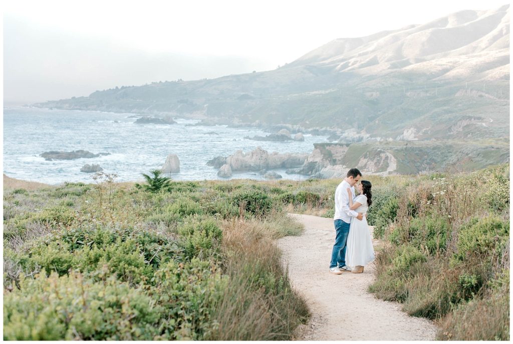 portrait of couple standing on a pathway with plants around them and the sea in the background