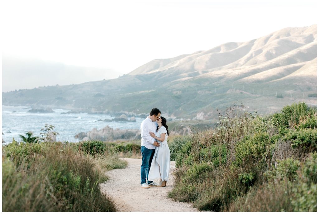 couple kissing surrounded by green plants with the ocean and hills behind them