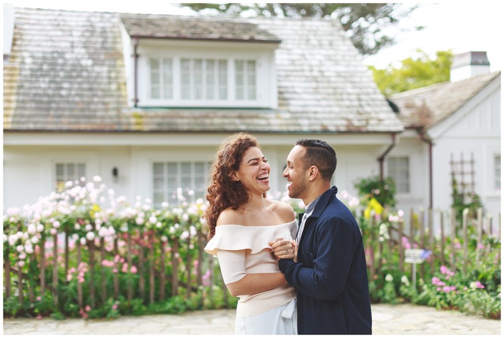 couple laughing in front of a vintage home with a picket fence and flowers