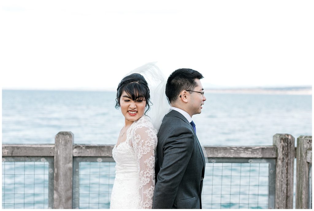 first look next to Monterey Bay back to back holding hands bride peaking at her groom asian couple