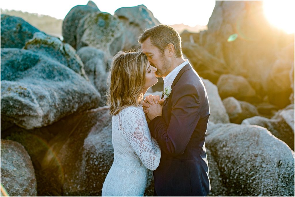 Couple kissing with a sun beam shining on them and beach rocks in the background.