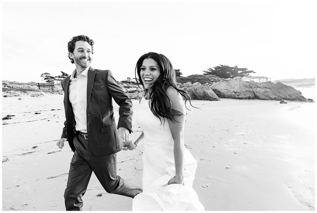 black and white photo of couple hand in hand smiling and running down Pebble Beach by film photographer Ags Photo Art