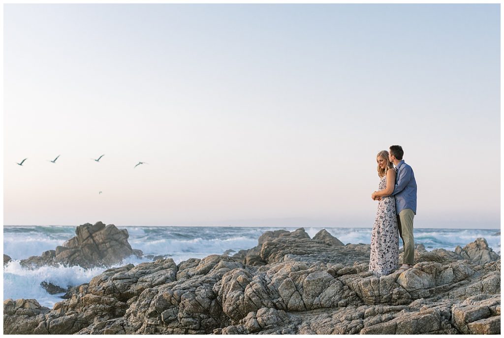 landscape photo of groom kissing his bride on her temple while they stand on the rocks of Pebble Beach with birds flying and waves crashing in the background by film photographer Ags Photo Art