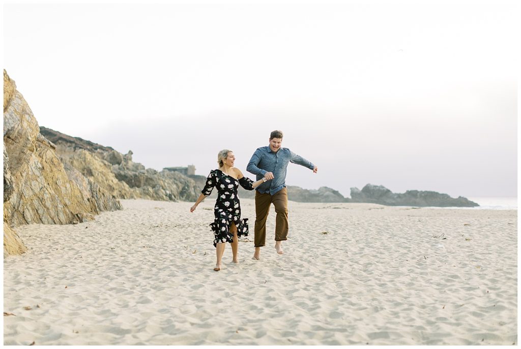 couple running and dancing joyfully in the sand Garrapoda State Park Beach in Big Sur, CA after their engagement at Folktale Winery