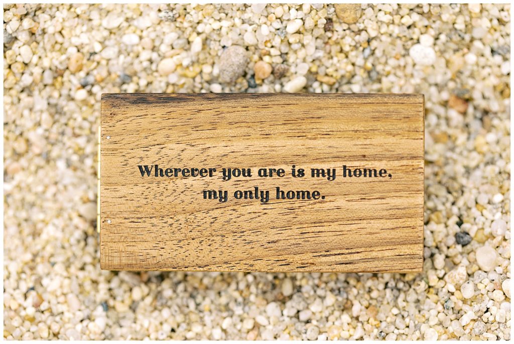 a wooden ring box on the pebbly sand for the California Coastline In N Out Surprise Proposal with the words "wherever you are is my home, my only home" engraved into it