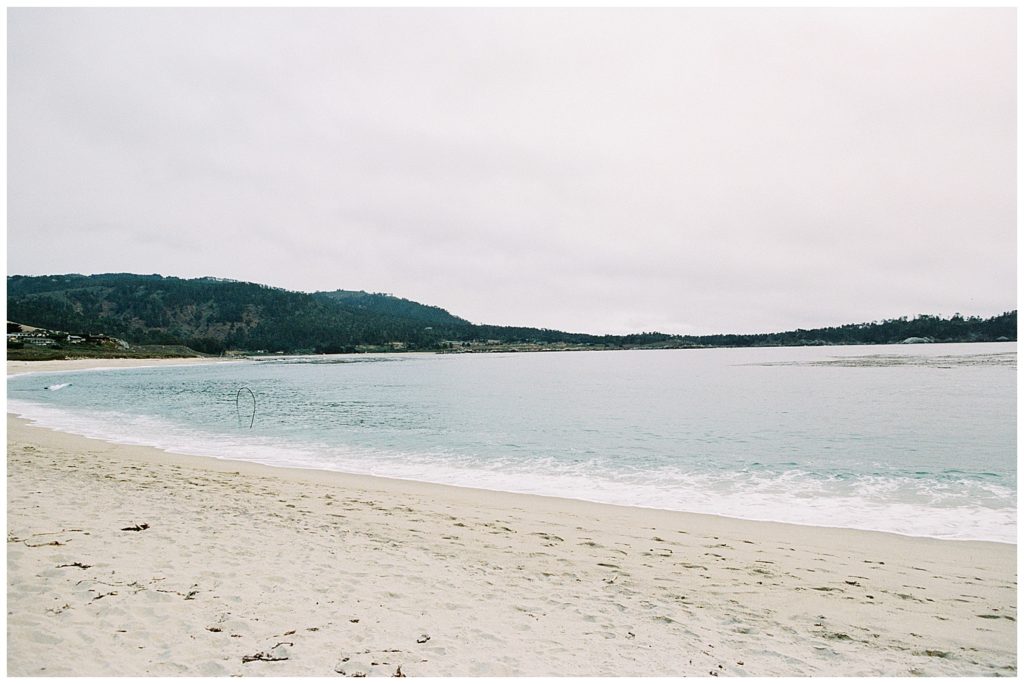 a landscape portrait of Carmel by the Sea beach under a light gray sky with soft blue and foamy white waves washing up on the shore by film photographer AGS Photo Art