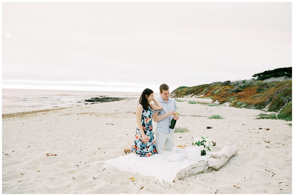 the couple kneeling down on their white picnic blanket on the California Coastline for their In N Out Surprise Proposal, the man is beginning to open the bottle of champagne while the woman watches him smiling