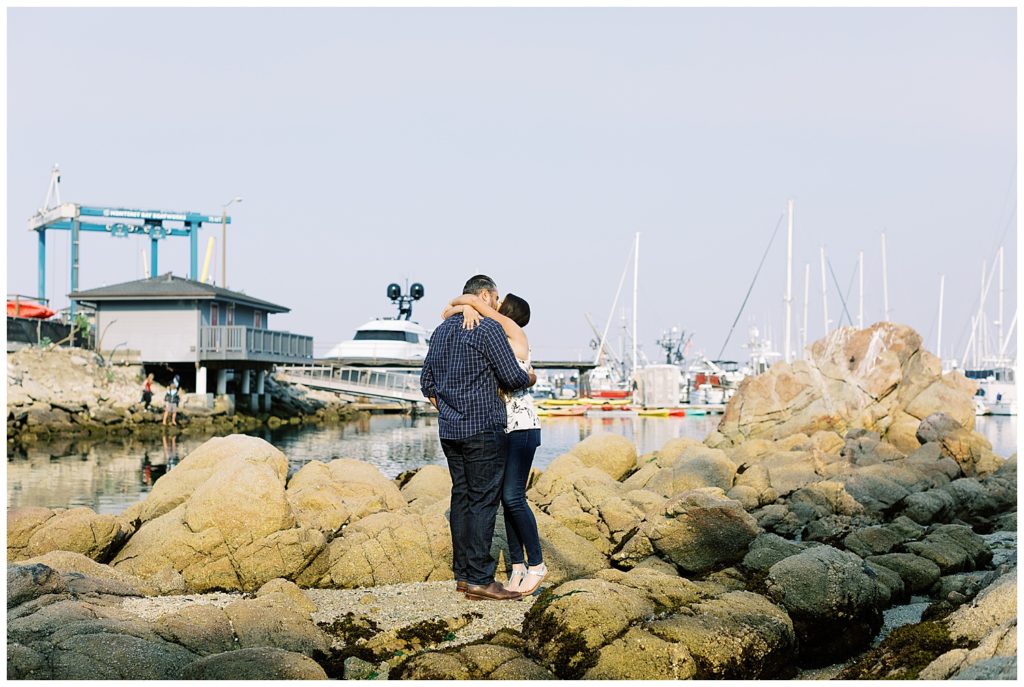 landscape portrait of the couple kissing on the rocks by the pier with boats in the harbor after their Beautiful And Whimsical Monterey Surprise Proposal by film photographer AGS Photo Art