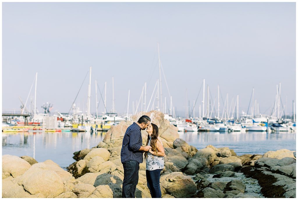 landscape portrait of couple during their Beautiful And Whimsical Monterey Surprise Proposal touching their noses together and holding hands with a rocky pier and many sailboats in the background by film photographer AGS Photo Art