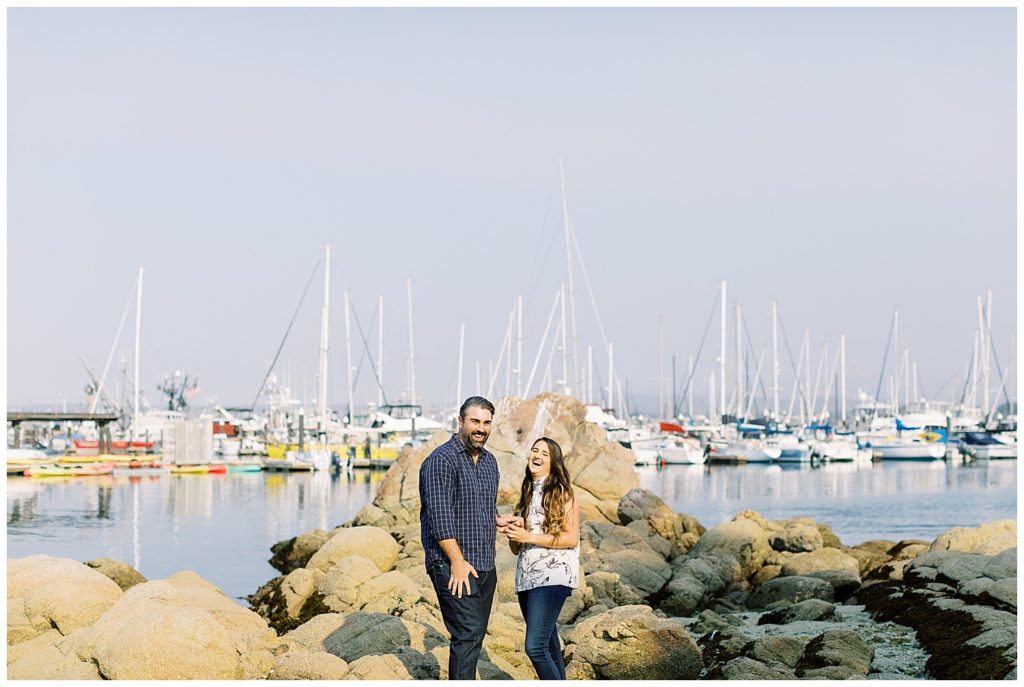 smiling and surprised woman holding her fiancé's hand after he proposed to her with many sailboats floating behind them in the background