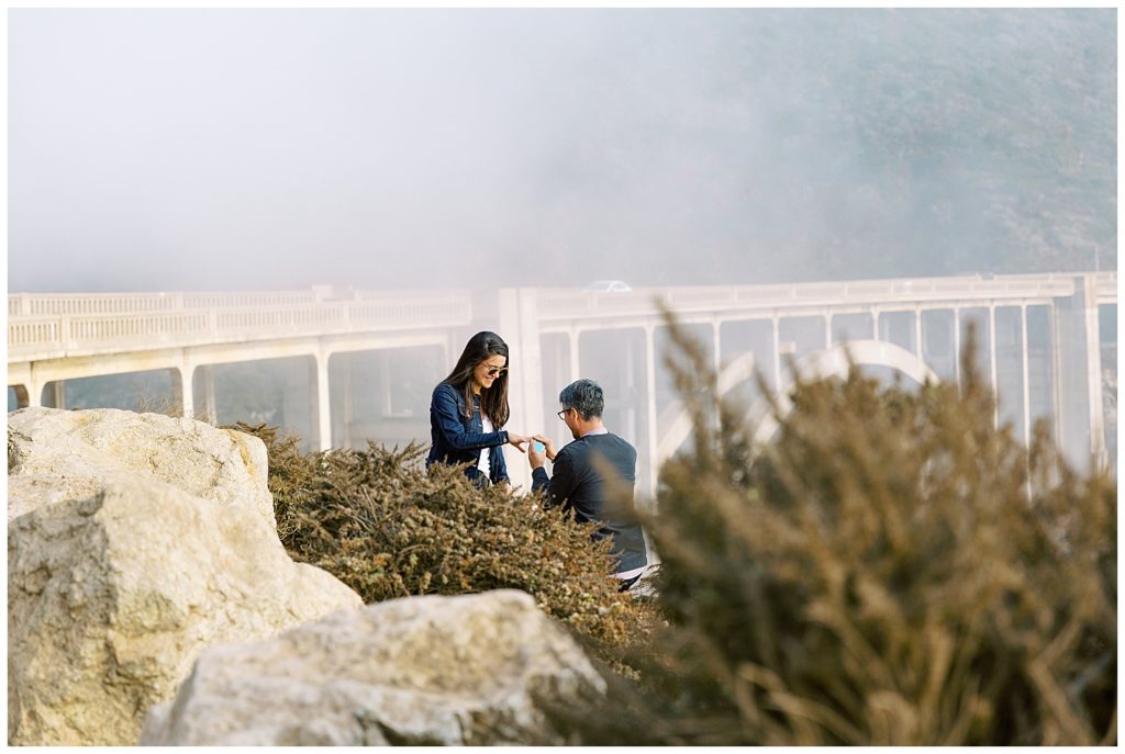 Beautiful Surprise Proposal At Bixby Bridge with Big Sur fog hovering over the bridge in the background as the man gets down on one knee to propose by film photographer AGS Photo Art