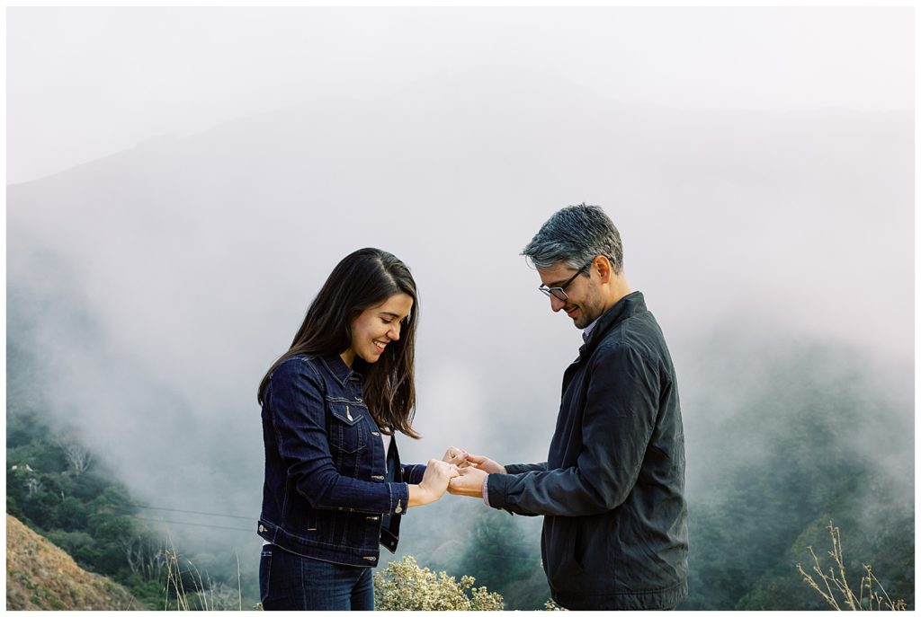 fog coming down the green Big Sur mountains as the couple looks at their rings from the surprise proposal