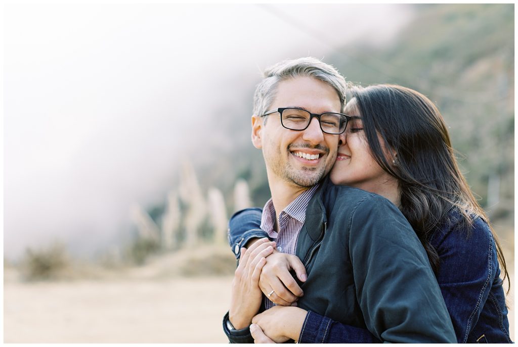 the couple joyfully embracing each other from behind with wide smiles on their faces and the foggy Big Sur mountains behind them