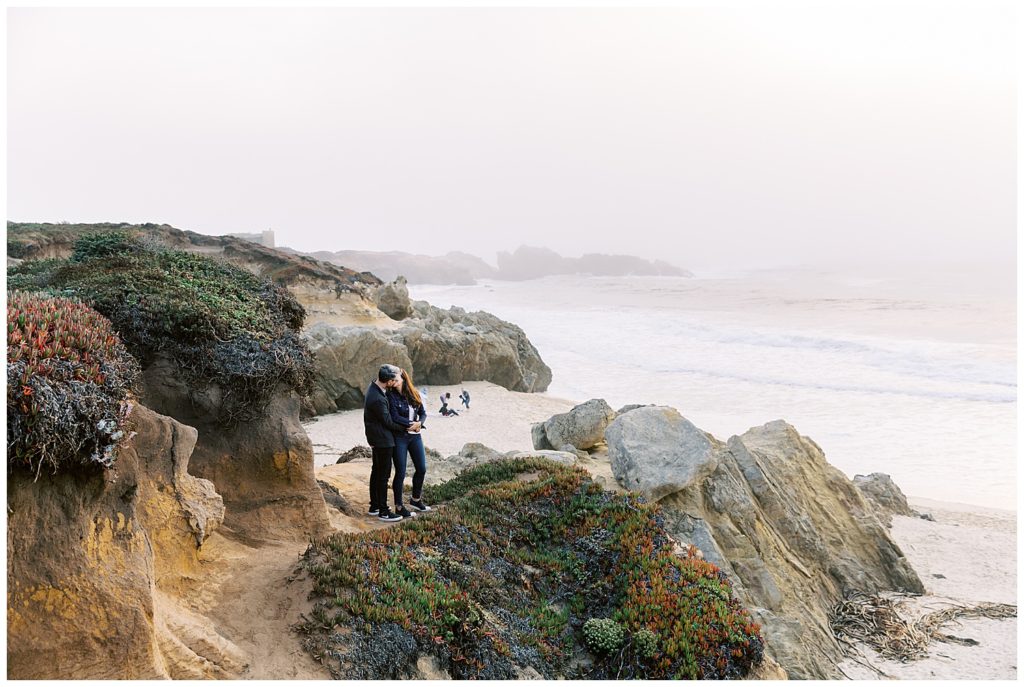 sweeping view of the green cliffs, white sand, and foamy waves of Big Sur, CA with the couple sharing a kiss after their surprise proposal at Bixby Bridge by film photographer AGS Photo Art