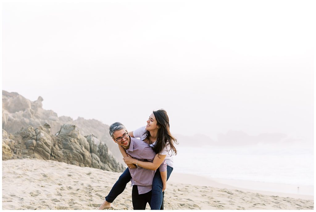 couple happily playing on the sands of a beach in Big Sur, CA the man gives his smiling fiancée a piggyback ride