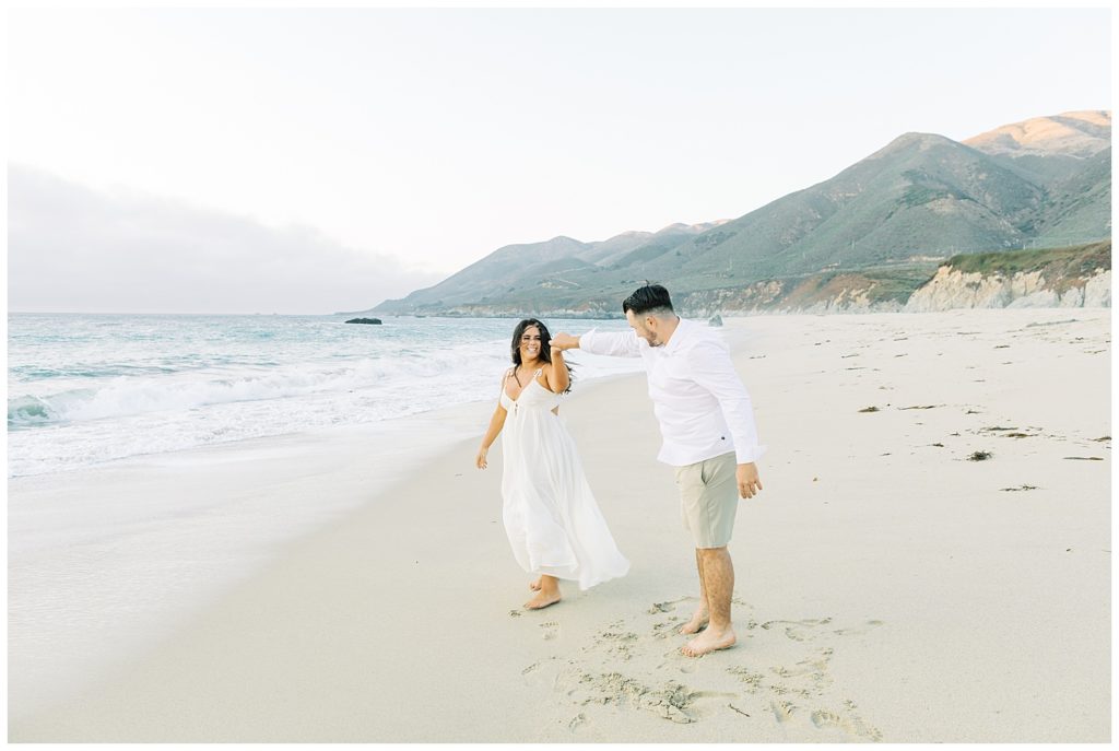 The man twirls his smiling fiancée on the sandy Big Sur shoreline by film photographer AGS Photo Art