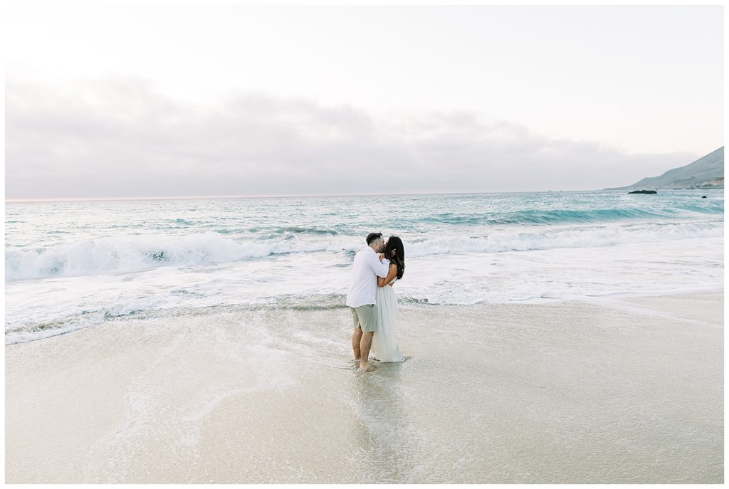 The couple shares a kiss during their lovely engagement in Beautiful Big Sur on the shoreline with blue and foamy white waves crashing in the background