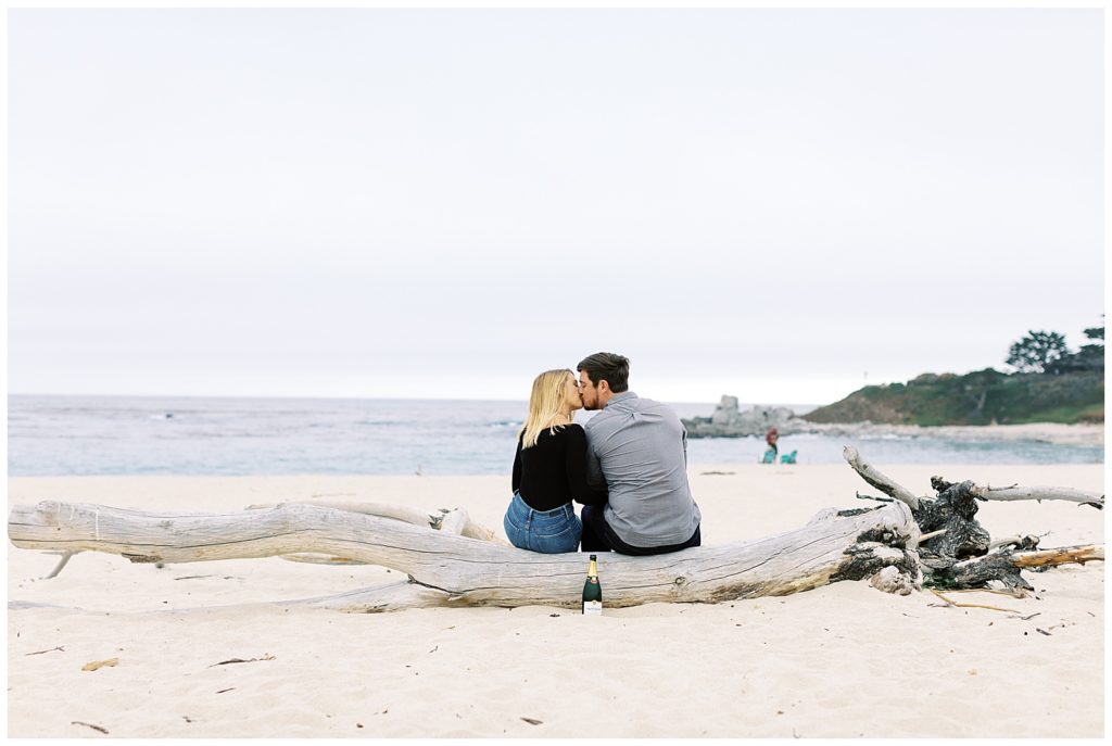 The couple sits on a driftwood log on the shore of Carmel Beach and shares a kiss to celebrate their Powerful And Heartfelt Anniversary Session by film photographer AGS Photo Art