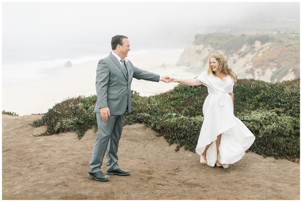 groom twirling his bride on their Big Sur elopement day on the sandy cliffs with the beach behind them by film photographer AGS Photo Art