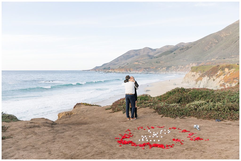 the couple embracing each other in celebration of their engagement in Big Sur surrounded by red rose petals forming a circle on the ground by film photographer AGS Photo Art