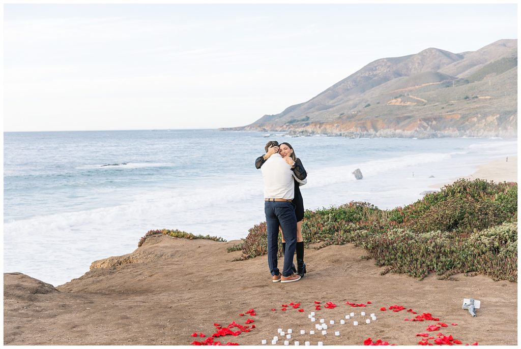 smiling couple in a tight hug celebrating their Big Sur proposal with the beach waves crashing in the background