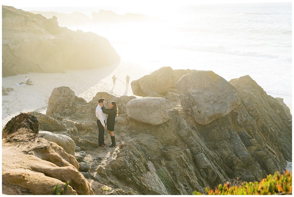 the woman holding her fiancé's face in her hands and smiling as they stand near the cliffs of the beach in Big Sur CA