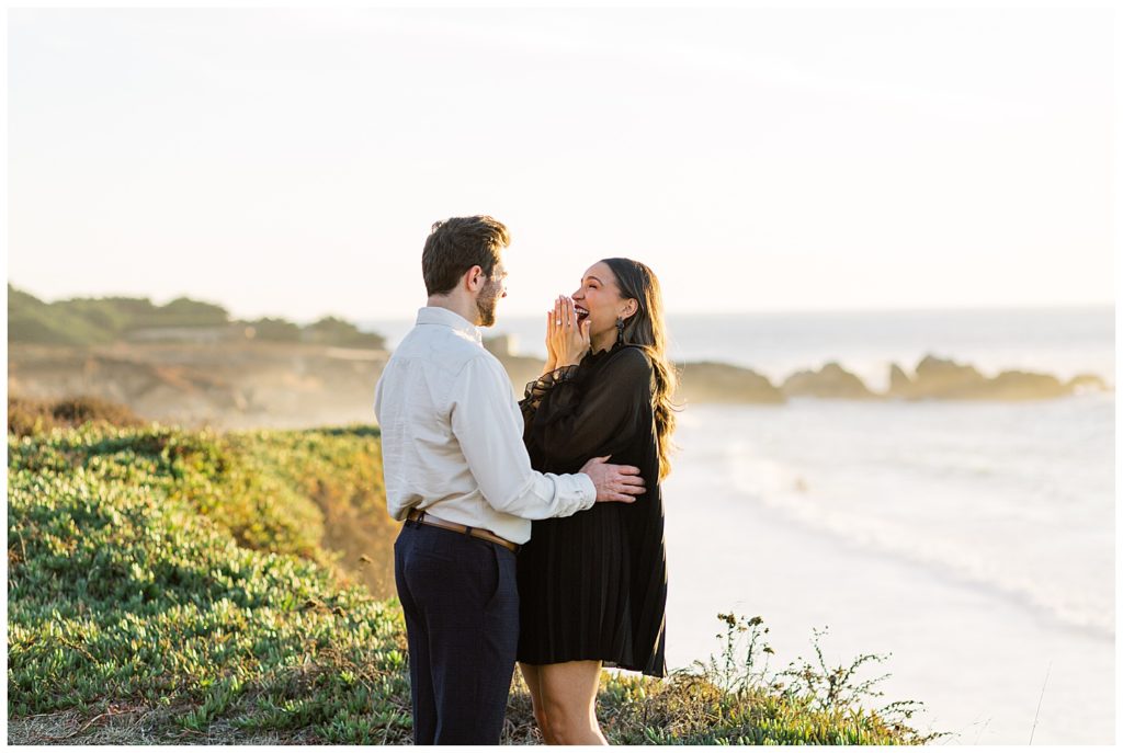 overjoyed woman grinning over her Surprise Proposal In Big Sur as her fiancé holds her close; the greenery and blue beach waves of Big Sur are lit up by the sunset in the background by film photographer AGS Photo Art