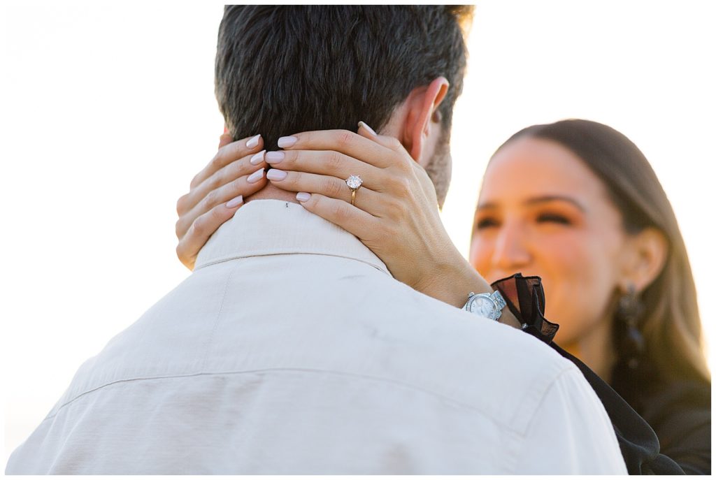 soft focus photograph of the woman's hands wrapping around her fiancé's neck, you can see her brilliant cut Surprise Proposal In Big Sur engagement ring on her pink manicure hands