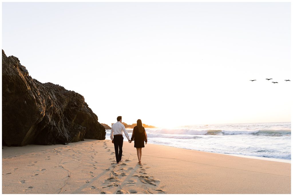 couple hand in hand walking down the beach in Big Sur away from the camera with the sun setting before them and birds flying overhead by film photographer AGS Photo Art