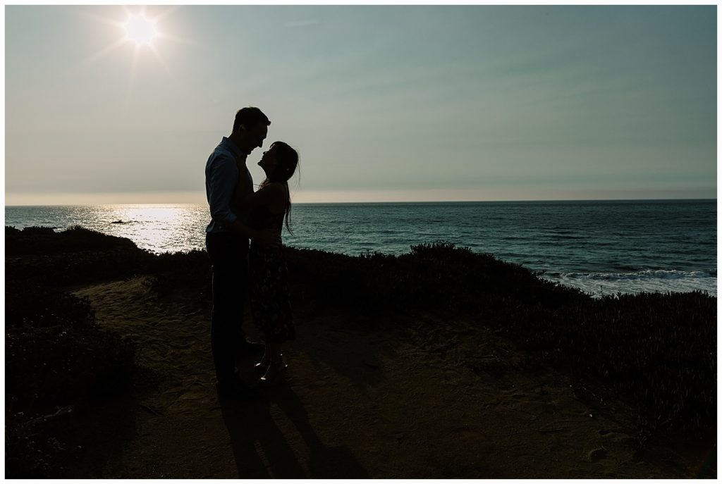 shadowed Big Sur Surprise Proposal portrait of couple smiling at one another with the sun shining overhead and the Pacific Ocean behind them by film photographer AGS Photo Art