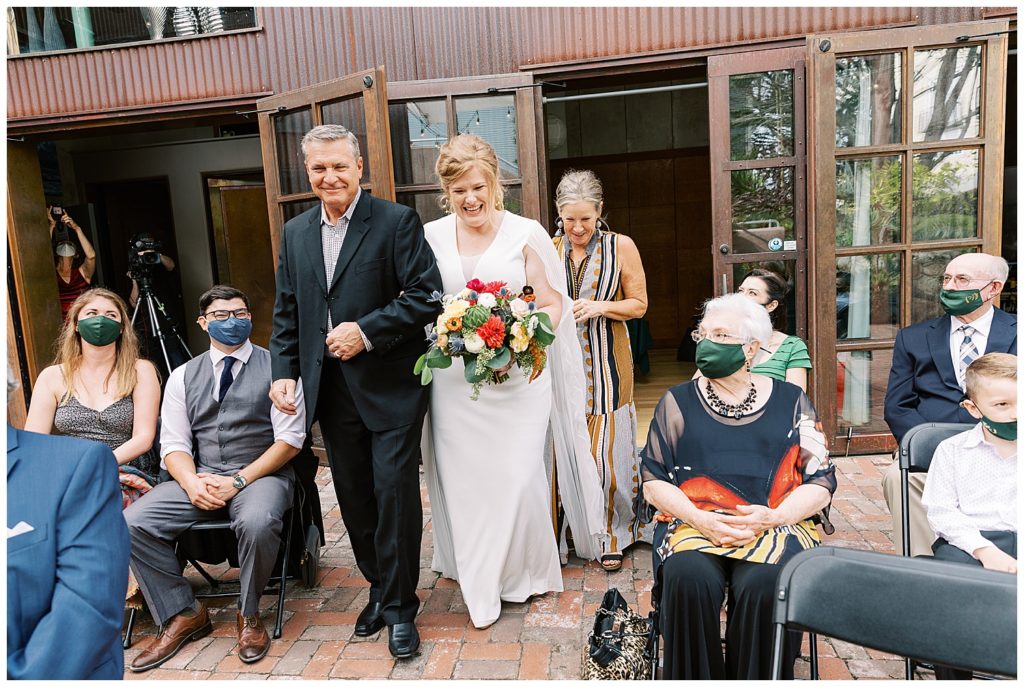 bride and parents walking down the aisle as their guests in masks watch them fondly