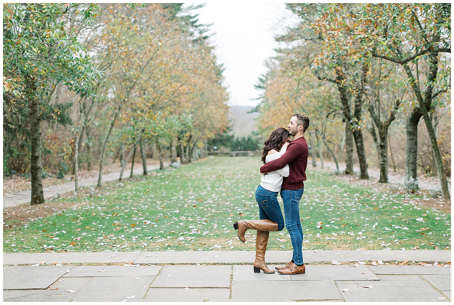 couple in New Jersey Botanical Garden for their engagement; the woman embraces her fiancé and tucks her leg up while he wraps his arms around her; they're both wearing brown shoes