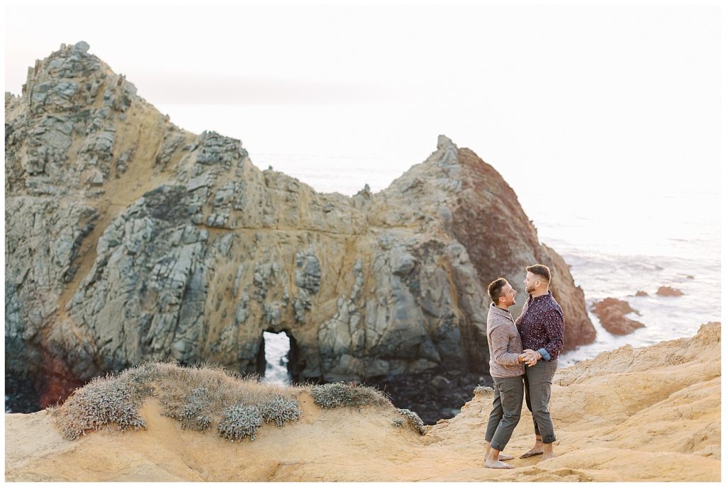 couple embracing each other and smiling as they stand on the cliffs of Pfeiffer Beach for their surprise sunset proposal by film photographer AGS Photo Art