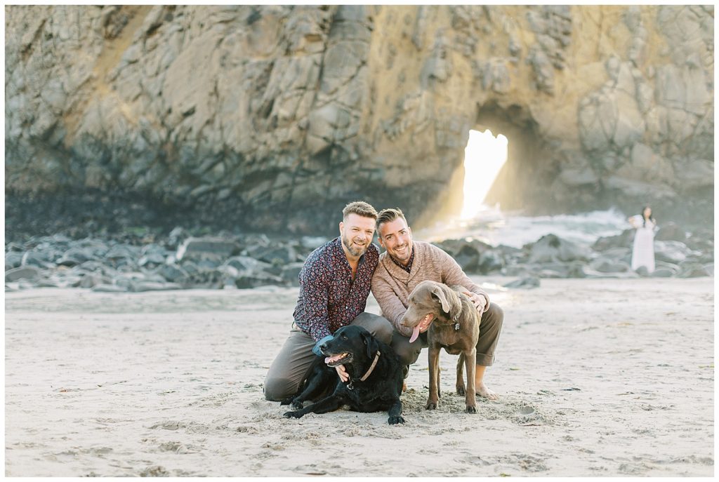 couple and their two labradors smiling at the camera at Pfeiffer Beach with sunshine pouring through the keyhole rock in the background