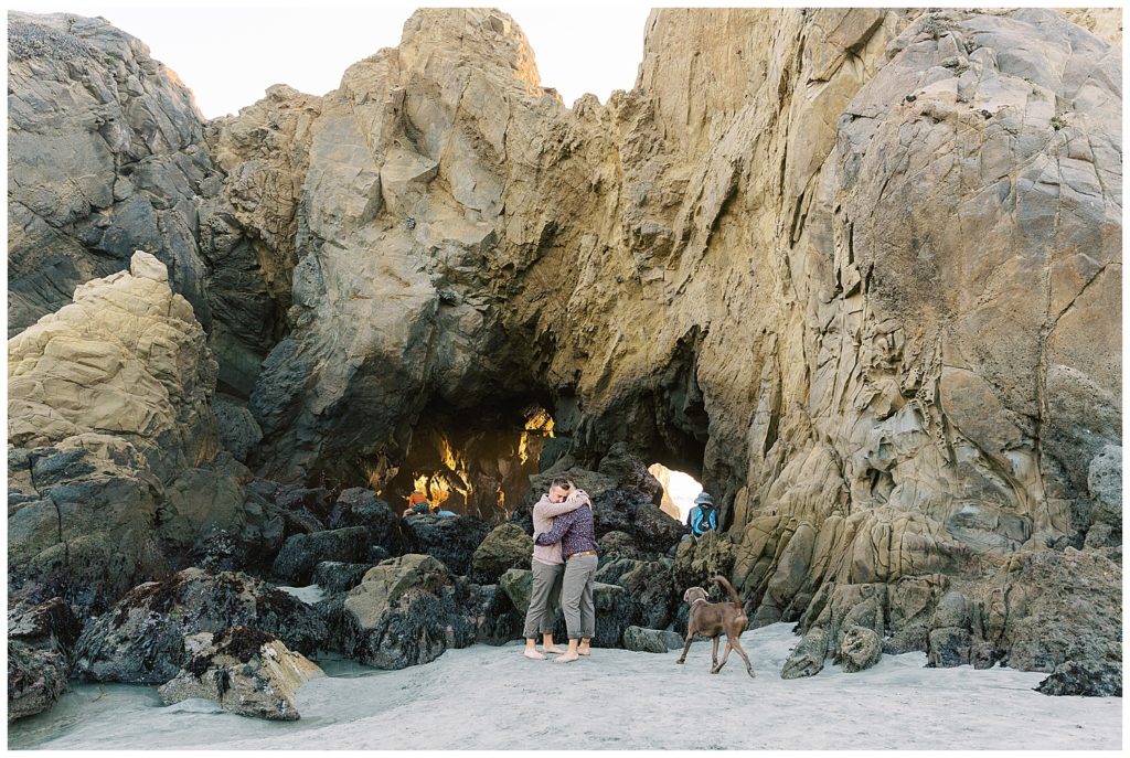 couple embracing at the foot of a large rock structure with one of their labradors running up to them
