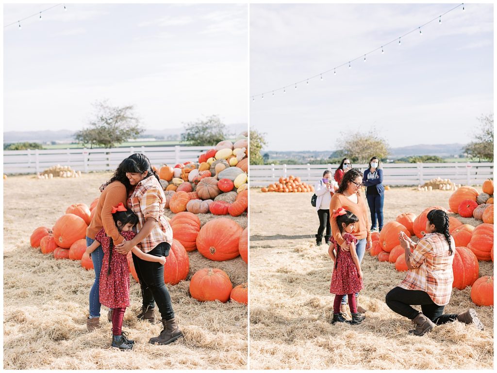 woman on one knee proposing in front of a pumpkin mountain to her fiancée with her daughter present