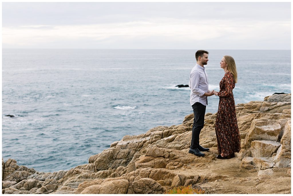 couple standing on a rocky cliff facing each other and smiling, their hands holding each other's while the Pacific Ocean rolls behind them