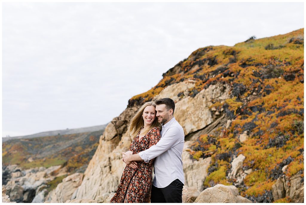 the man embracing his fiancée from behind as they smile at each other with orange and green grassy rock cliffs behind them in their Big Sur Sunset Engagement Shoot