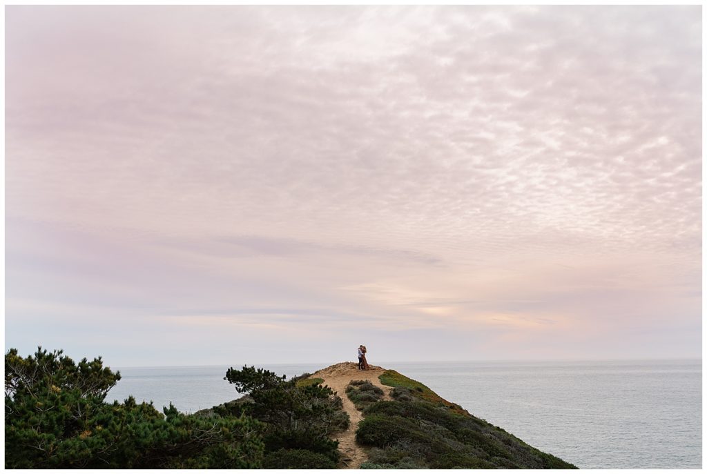 Cotton Candy Sunset Skies above the couple embracing on the top of a grassy cliff in Big Sur for their Sunset Engagement Shoot by film photographer AGS Photo Art