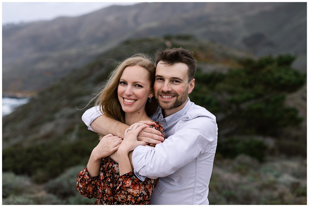 Big Sur Sunset Engagement Shoot portrait of the couple with the man's arms wrapped around his fiancée's chest while they both smile at the camera