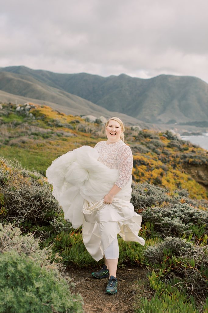 portrait of the bride during her Pebble Beach elopement; she's lifting up her wedding dress by BHDN to reveal comfortable hiking shoes underneath while she smiles at the camera