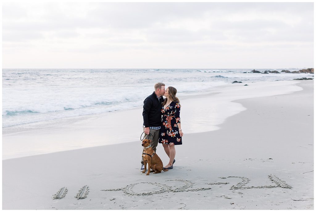 couple sharing a kiss at Pebble Beach for their engagement with their dog, Kona, sitting at their feet and their wedding date written in the sane