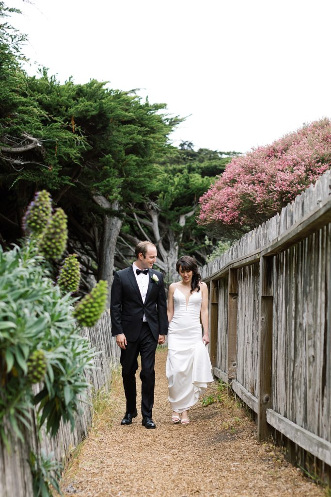 bride and groom hand in hand walking to the camera in between two wooden fences and greenery