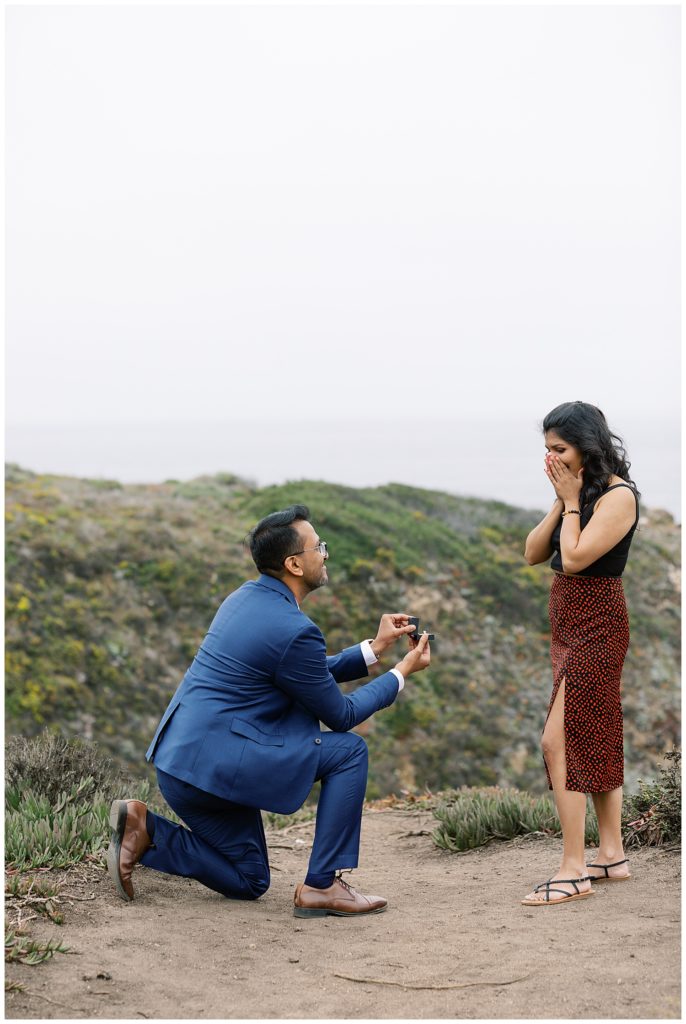 man in a blue suit popping the question in Big Sur to his now fiancée in a red and black outfit