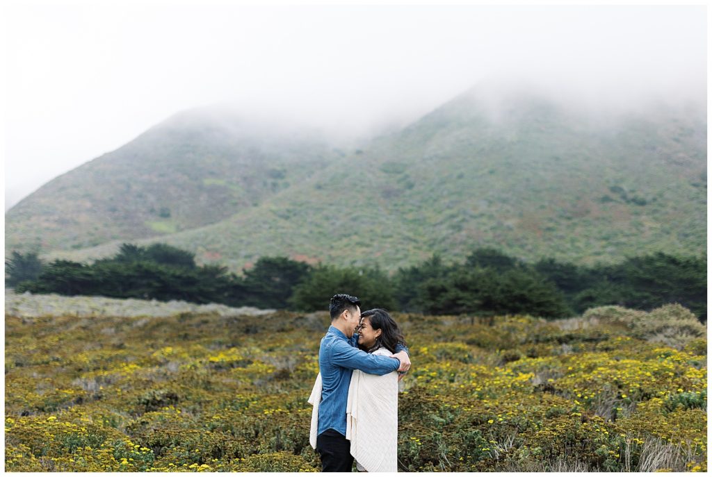 photo of the couple embracing in a flower field in Big Sur; the woman is wrapped in a white blanket and her fiancé's arms